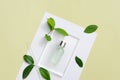 The labelless, transparent serum bottle is placed on a glass platform with fresh green tea leaves and a pastel background.