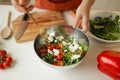 Woman putting cut feta cheese into bowl with tasty salad on table in kitchen and light blur background. Royalty Free Stock Photo