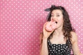 Lady with crezily pleased eyes is biting off a piece of doughnut with pink icing, while standing on pink background. Royalty Free Stock Photo