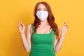 Lady with fingers crossed wishing best against yellow background, redhaired ginger woman feeling unhappy and wearing medical mask