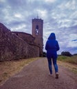 A lady with her back turned wearing a blue hooded coat walking towards a church under cloudy sky in Monells Royalty Free Stock Photo