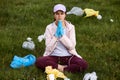 Lady sitting on ground in green meadow and praying for better for environment, keeping hands together surrounded with trash, being