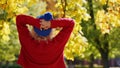 Laid-back caucasian young person standing backwards to the camera in red sweater and blue winter hat. Autumn colors