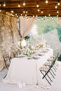 Laid festive table with chairs stands against a stone wall under a wooden canopy with glowing garlands
