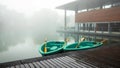 A lake with a cafe building near the water in the fog. In the foreground, a pier with paddle boats wet from rain. Royalty Free Stock Photo