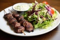 lamb kebabs on white plate, mixed salad on the side