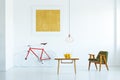 Lamp above wooden table in white flat interior with green armchair and yellow painting above bike. Real photo