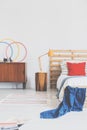 Lamp on wooden stool next to bed with red cushion and blue blanket in bedroom interior. Real photo