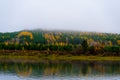 The landscape of the autumn riverbank with hills covered with dense forest of green and yellow leaves. Fog over the taiga river in