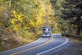 Landscape with blue big rig semi truck and bulk semi trailer running on the winding road with autumn forest on the sides Royalty Free Stock Photo