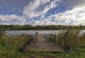 Landscape with wooden long jetty with chair for fishing, lake, forest on horizon and cloudy blue sky in summer Royalty Free Stock Photo