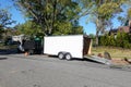 A landscaping truck with a long white enclosed trailer trailer with it`s rear ramp down seen on a shady residential asphalt stree