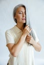 Languid middle aged Asian woman strokes lock of silver hair on light grey background