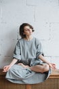 Languid young woman in grey dress sitting in half lotus pose on a low cupboard