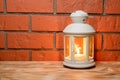 A lantern with a glowing candle in on the wooden table, brick wall background