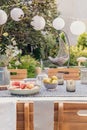 Lanterns above table with food and drink in the garden with plants and wooden chairs. Real photo