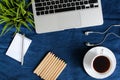 Laptop keyboard, white cup of tea on saucer, notepad, pen and green plant in the corner on dark blue crumpled jeans background. Royalty Free Stock Photo