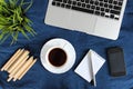 Laptop keyboard, white cup of tea on saucer, notepad, pen and green plant in the corner on dark blue crumpled jeans background. Royalty Free Stock Photo