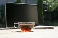 Laptop, phone and cup of tea with shining ray of sun light in it on light wooden table outside in green summer garden. Royalty Free Stock Photo