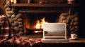 Laptop with a picture of a bonfire screen, indoor near burning fireplace in rustic style, with cozy blanket and cup of coffee.