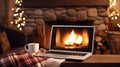 Laptop with a picture of a bonfire screen, indoor near burning fireplace in rustic style, with cozy blanket and cup of coffee.