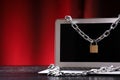 Laptop tied with an iron chain with a lock on a dark office table against black and red background. Cyber security and cyber crime