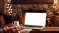 Laptop with a white screen mock up, indoor near burning fireplace in rustic style, with cozy blanket and cup of coffee. Seasonal