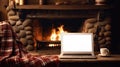 Laptop with a white screen mock up, indoor near burning fireplace in rustic style, with cozy blanket and cup of coffee. Seasonal