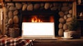 Laptop with a white screen mock up, indoor near burning fireplace in rustic style, with cozy blanket and cup of coffee. Seasonal