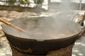 Large black cauldron on the street in which a brown food is cooked and there is steam