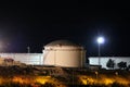 Large brightly lit white industrial oil refinery tanks with protective fence placed on top of small hill on warm summer night