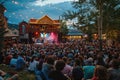A large crowd of people eagerly sits in front of a stage, waiting for an exciting performance to begin, A bustling outdoor music