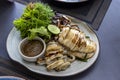 A large grilled squid looks appetizing with a small cup of spicy sauce next to it, served with vegetables in a white porcelain