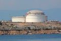 Large industrial white oil refinery tanks with protective fence placed on top of small hill surrounded with rocky shore and calm