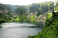 A large lake in a mountain hollow with snow on the slopes and a dense overgrowth of bushes under a cloudy summer sky