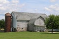 A large old stone foundation white barn and stone foundation and brick silo with a bright green grass lawn and shade trees