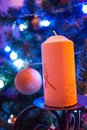 A large orange candle on a wrought-iron stand against the background of a Christmas tree decorated with toys is a detail