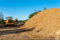 A large pile of wood chips lying on a square, with a blue sky in the background, visible bulldozer