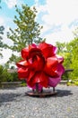 A large red and pink metal flower in the garden surrounded by lush green trees and plants with blue sky and clouds