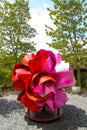 A large red and pink metal flower in the garden surrounded by lush green trees and plants with blue sky and clouds