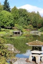 A large reflecting pond with a tourist taking photos and a variety of evergreen, Japanese Maples and Willow trees