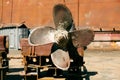 Large, rusted ship propeller displayed on wooden blocks in a shipping yard, symbolizing industrial maritime decommission