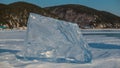 A large shiny ice floe stands vertically on a frozen and snow-covered lake.