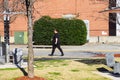 A large size man dressed in black walking along a sidewalk in front of a red brick buildings surrounded by bare winter trees