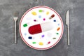 Large sized red pill on a plate with silverware, in the middle of colorful medicine on grey slate background Royalty Free Stock Photo
