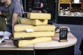 Large slabs of cheese on wooden table at a market with worker leaning over talking to customer and food allergies sign on tables