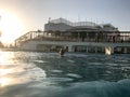 A large stone building with a pedestrian bridge with arches above the pool near the water, a pool against a blue sky and a big sun Royalty Free Stock Photo