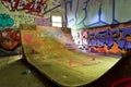 A large sunlit plywood skateboard ramp surrounded by walls covered in bright graffiti.
