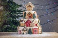 Large tiered Christmas cake decorated with gingerbread cookies and a house on top. Tree and garlands in the background.