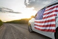 large US flag is affixed to side of car driving down country road through rapeseed fields at sunset Royalty Free Stock Photo